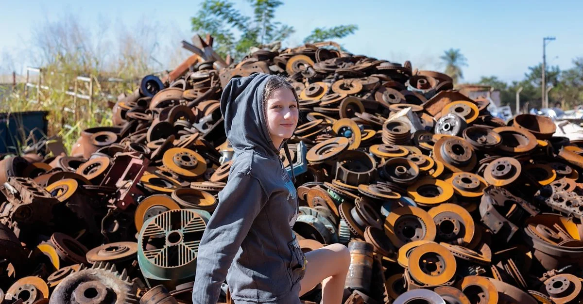 Une adolescente en hoodie pose avec confiance à côté d'un grand tas de ferraille rouillée en extérieur.